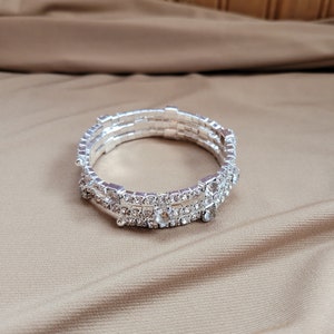 Puede incluir: Un brazalete plateado con tres filas de diamantes de imitación transparentes. El brazalete está sobre un fondo de tela marrón.
