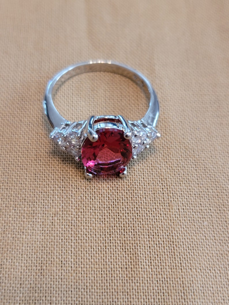 May include: A silver ring with a large, oval-shaped, pink gemstone surrounded by smaller, clear gemstones.