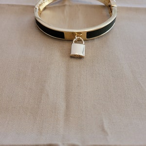 May include: Gold-tone hinged bracelet with a black enamel band and a small, silver-coloured padlock charm. The bracelet has a simple, elegant design. The padlock charm adds a unique touch. Suitable for everyday wear or special occasions.
