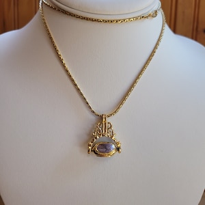 May include: A gold chain necklace with a pendant featuring a purple stone set in a gold frame. The pendant is shaped like a small archway with intricate details.