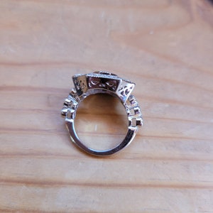 May include: A silver ring with a large, square-shaped stone set in the center. The ring is decorated with intricate details around the stone and band.