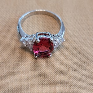 May include: A silver ring with a large oval-shaped red gemstone surrounded by smaller clear gemstones.