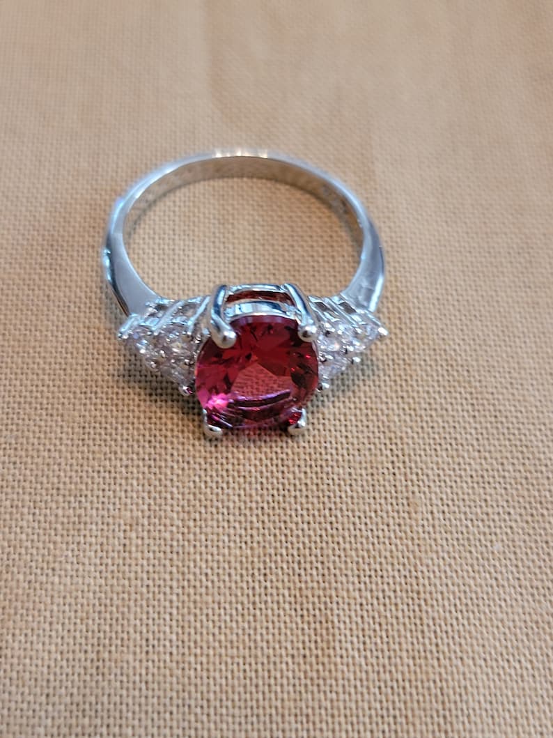 May include: A silver ring with a large oval-shaped red gemstone surrounded by smaller clear gemstones.