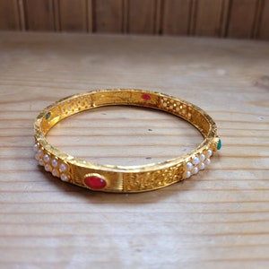 May include: A gold bangle bracelet with a decorative design and white pearl accents. The bracelet has red and green gemstones.
