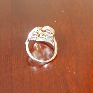 May include: A silver ring with a heart-shaped design and a clear stone in the center. The stone is surrounded by smaller stones.