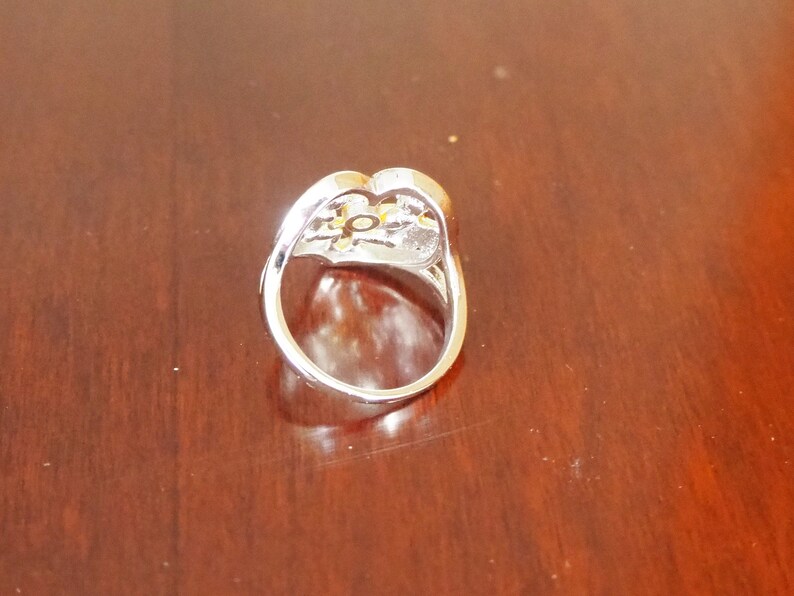 May include: A silver ring with a heart-shaped design and a clear stone in the center. The stone is surrounded by smaller stones.