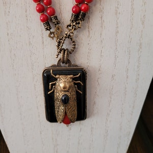 May include: A red coral beaded necklace with a gold-toned pendant featuring a black and gold cicada. The pendant has a black background with a gold cicada with a black gemstone in its head.