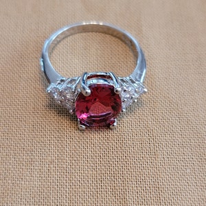 May include: A silver ring with a large, oval-shaped, red gemstone surrounded by smaller, clear gemstones.