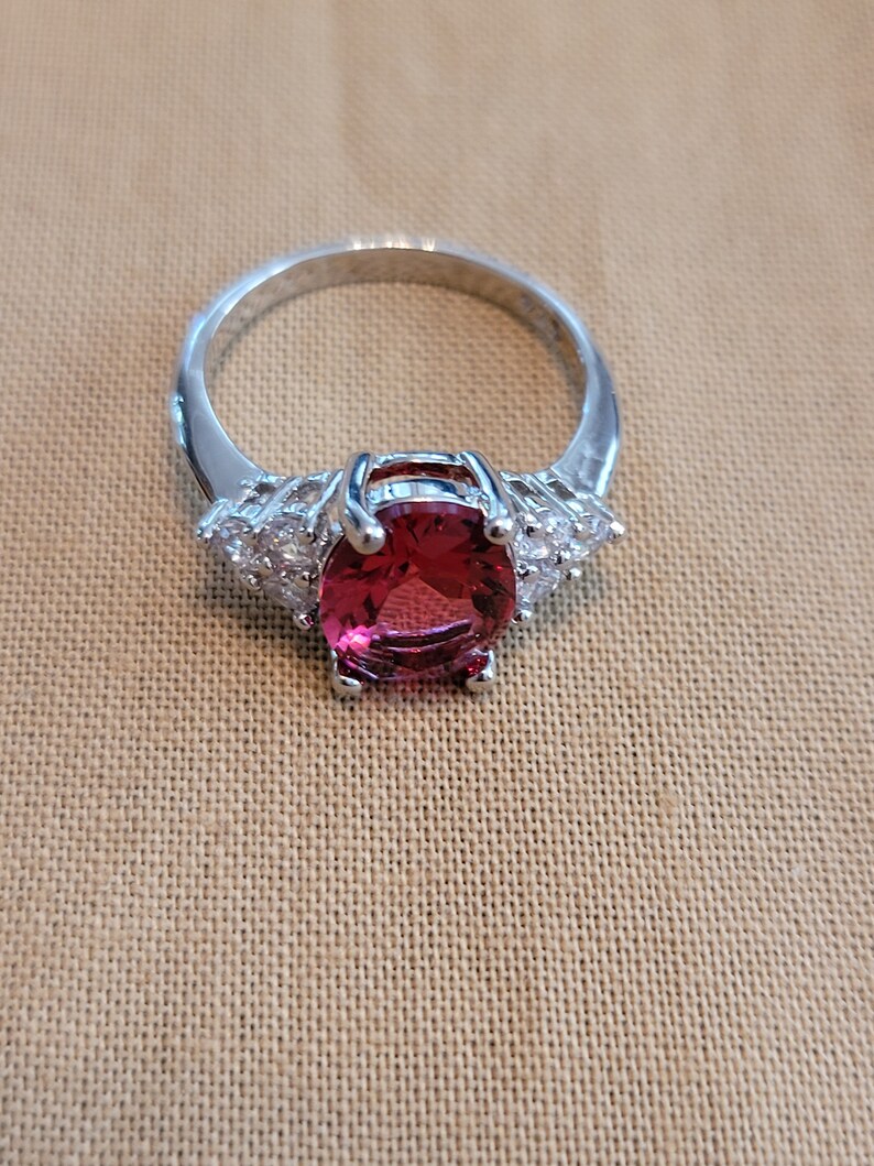 May include: A silver ring with a large, oval-shaped, red gemstone surrounded by smaller, clear gemstones.