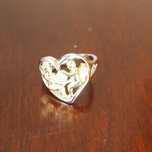 May include: A silver ring with a heart-shaped design featuring two gold angels holding a small clear stone. The ring is on a wooden surface.