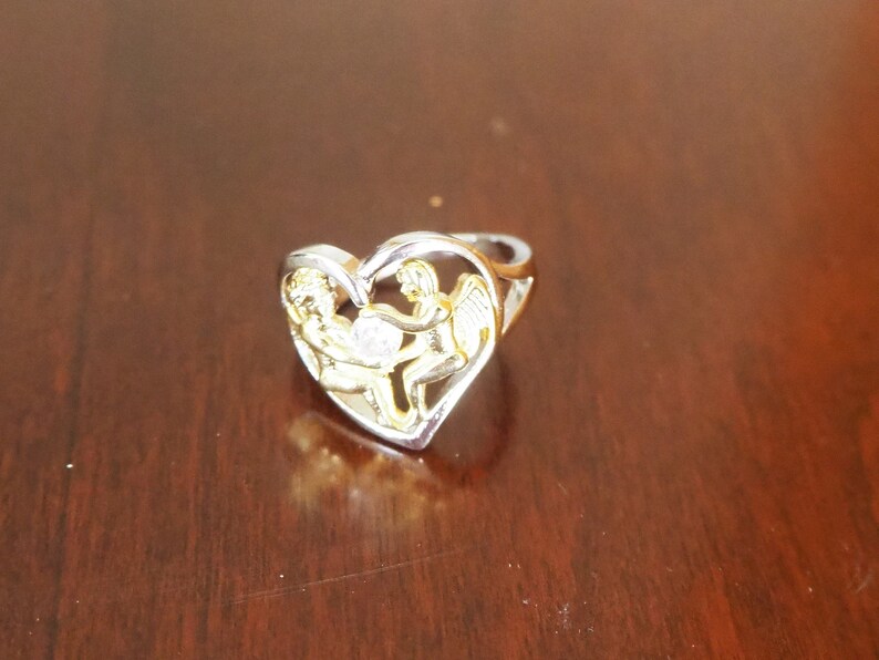 May include: A silver ring with a heart-shaped design featuring two gold angels holding a small clear stone. The ring is on a wooden surface.