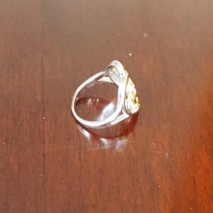 May include: A silver ring with a gold-toned design in the shape of a heart. The ring is on a wooden surface.