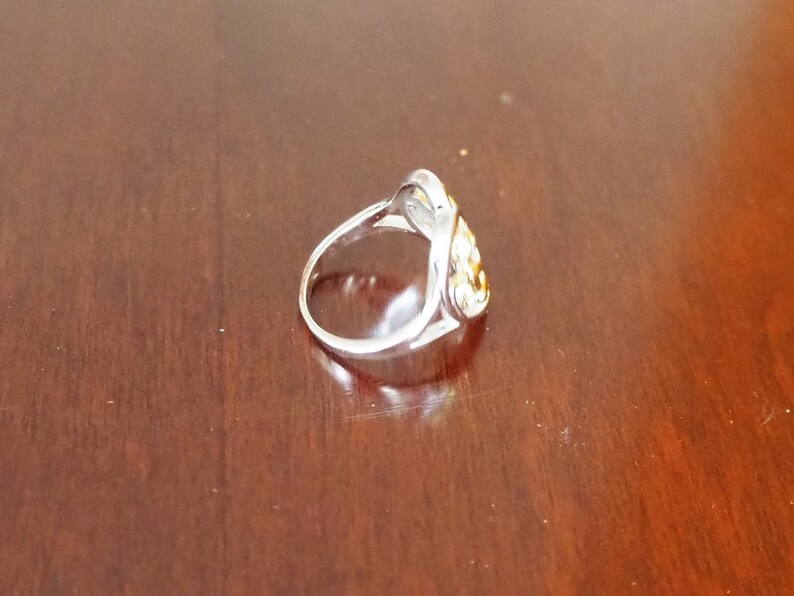 May include: A silver ring with a gold-toned design in the shape of a heart. The ring is on a wooden surface.
