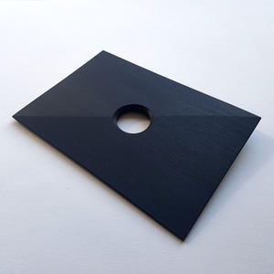 Könnte beinhalten: Eine schwarze rechteckige Holzplakette mit einem runden Loch in der Mitte. Die Plakette hat eine Holzmaserung.