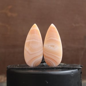 Pode incluir: Dois cabochões de ágata em forma de lágrima com um padrão de faixas brancas e laranja. Os cabochões são polidos e têm um acabamento liso e brilhante.