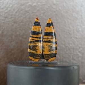 May include: Pair of teardrop-shaped gemstone cabochons with a tiger eye pattern in shades of brown, yellow, and black. The cabochons are polished and have a smooth, glossy finish.