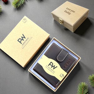 May include: A dark brown leather wallet with white stitching, presented in a gift box. The wallet is in a box with the brand name "PW" and the words "U DAD" embossed on it. A wooden box with "Love you Daddy" is also present.