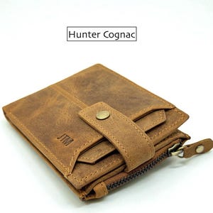 Könnte beinhalten: Eine braune Lederbrieftasche mit Druckknopfverschluss und Reißverschluss. Die Brieftasche hat ein Used-Finish und der Text "Hunter Cognac" steht darüber. Die Initialen "JTM" sind eingeprägt.