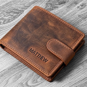Op de afbeelding: Een bruine leren bifold portemonnee met een verweerde afwerking en een veilige sluiting met drukknop. De naam MATTHEW is in hoofdletters in de voorkant van de portemonnee gedrukt, die langs de randen is voorzien van zichtbare stiksels.