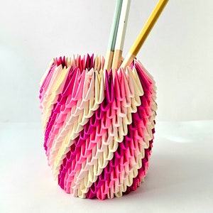 May include: A pink and white origami paper vase with two wooden chopsticks inside. The vase is made of folded paper and has a unique, textured design.