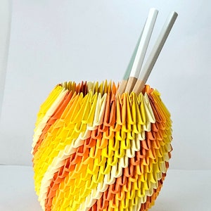 May include: A yellow, orange, and white origami vase with two white chopsticks inside.