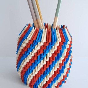 May include: A cylindrical pencil holder constructed from folded paper units in a repeating pattern of red, white, and blue. The structure features a textured, layered surface and contains four wooden pencils with colored tips in shades of gray, yellow, and green.