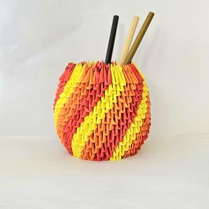 May include: A colorful, handmade origami pen holder in shades of yellow, orange, and red. The spherical container holds three pencils: black, light brown, and tan. The origami design creates a textured, geometric pattern.