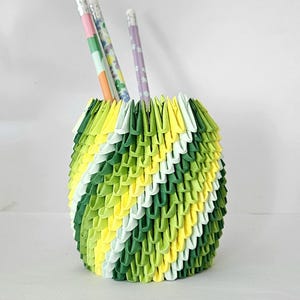 May include: A handmade origami pencil holder in shades of green, yellow, and white, with three pencils inside. The holder has a textured, geometric design. The pencils have pink, green, and purple accents.