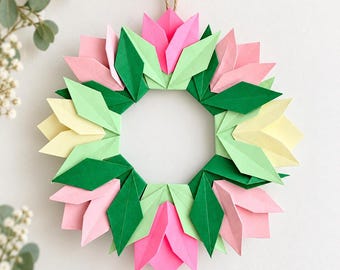 May include: A circular wreath constructed from folded paper elements in shades of hot pink, light pink, pale yellow, light green, and dark green. The geometric petals and leaves are arranged in a repeating pattern, suspended by a tan twine loop.