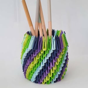 May include: A colorful, handmade origami pen holder. The holder is a cylinder shape, with a layered design in shades of green, blue, and purple. Several pencils are visible inside the holder, with their wooden bodies and colored tips.