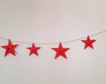 May include: A red paper star garland with six stars hanging on a white string. The stars are made of folded paper and have a three-dimensional look.