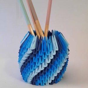 May include: A blue and white origami pencil holder with three pencils sticking out of the top. The pencils are painted in different colors: light blue, pink, and brown.