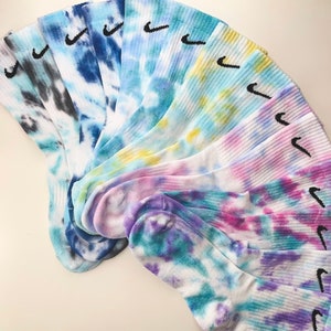 Può includere: Una collezione di calzini tie-dye in vari colori, tra cui blu, viola e giallo. Ogni calzino presenta il logo Nike swoosh in nero. I calzini sono disposti a ventaglio, mostrando i motivi colorati.