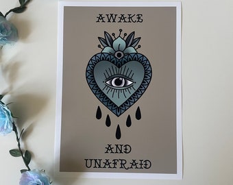 May include: A framed print featuring a teal heart with an eye, floral accents, and teardrops. The words "AWAKE" and "AND UNAFRAID" are printed above and below the heart. The background is a muted tan color.