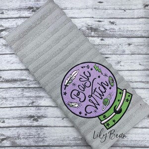 May include: A grey kitchen towel with an embroidered design of a purple crystal ball with the text "Basic Witch" in green and purple lettering. The design also includes stars, a crescent moon, a feather, and a lightning bolt. The text "Lily Bean" is embroidered in the bottom right corner.