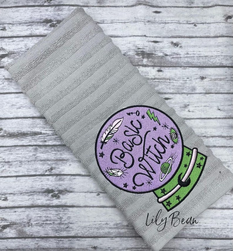 May include: A grey kitchen towel with an embroidered design of a purple crystal ball with the text "Basic Witch" in green and purple lettering. The design also includes stars, a crescent moon, a feather, and a lightning bolt. The text "Lily Bean" is embroidered in the bottom right corner.