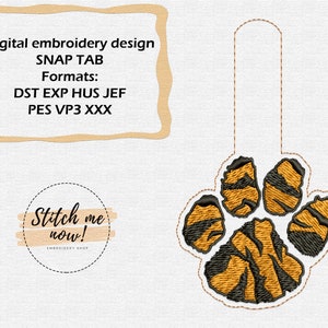 Op de afbeelding: Tigerpootafdruk borduurontwerp voor een drukknoop. Het ontwerp is oranje en zwart met een gestreept patroon. De tekst "Stitch me now!" staat in een cirkel onder de poot.