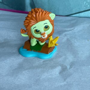 May include: A small, cartoon lion figurine with a green body, orange mane, and a blue base. The lion has large eyes and a friendly expression. It is standing on a brown and green platform with a yellow flower.