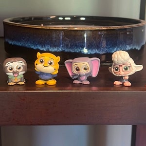 May include: A set of six small, colorful figurines of characters from the animated movie Zootopia. The figurines are made of plastic and feature the characters Judy Hopps, Nick Wilde, Flash, Finnick, Mr. Big, and Bellwether.
