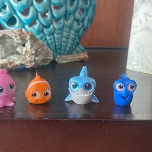 May include: Five small plastic figurines of characters from the movie Finding Nemo. The characters are a pink octopus, an orange clownfish, a blue shark, a blue tang, and a green sea turtle.
