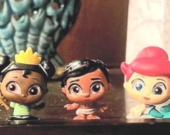 May include: A set of six small, colorful figurines of Disney princesses. The figurines are made of plastic and have a cartoonish style. The princesses include Ariel, Tiana, Moana, Mulan, Pocahontas, and Rapunzel.
