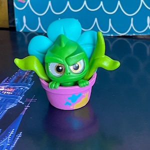 May include: A green cartoon character with a grumpy expression, resembling a plant, sits in a pink flower pot. The character has large eyes and is surrounded by teal and green leaves. The pot has blue and yellow accents.