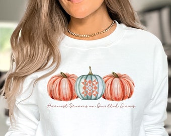 May include: White sweatshirt featuring a watercolor design of three pumpkins in shades of orange, teal, and coral. The center pumpkin has a quilt-inspired pattern. The text "Harvest Dreams on Quilted Seams" is below the pumpkins.