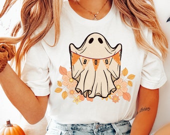 May include: White t-shirt featuring a cartoon ghost with a "BOO" banner and floral accents. The ghost design is in beige and black, with orange and peach flowers. The shirt is part of a Halloween-themed collection.