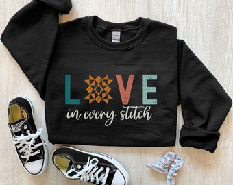 May include: Black sweatshirt with the text "LOVE in every stitch" in teal, orange, and white. The word "LOVE" has a quilt block design. Black Converse sneakers and a scrunchie are next to the sweatshirt.