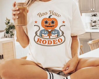 May include: Cream-colored t-shirt featuring a pumpkin head wearing sunglasses and a cowboy shirt. The shirt has the text "Boo Haw Rodeo" in a vintage style. The design includes stars and a rope border, perfect for Halloween or a rodeo theme.