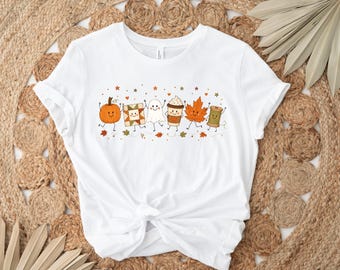 May include: A white short-sleeve t-shirt featuring a central graphic of six anthropomorphic characters including a pumpkin, a quilt block, a ghost, a coffee cup, an autumn leaf, and a spool of thread, all surrounded by small stars and leaves.