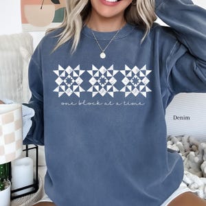 May include: A blue sweatshirt with a white design of three quilt blocks and the text "one block at a time".
