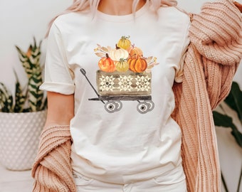 May include: Cream-colored t-shirt featuring a watercolor illustration of a wooden wagon filled with pumpkins. The wagon has a geometric pattern, and the pumpkins are in shades of orange, yellow, and white. The shirt is a casual style.