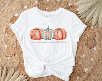 May include: White t-shirt featuring a watercolor design of three pumpkins. Two are orange, and one is teal with a quilt pattern. The text "Harvest Dreams on Quilted Seams" is printed below the pumpkins. The shirt is knotted at the waist.